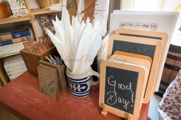 A photo of a desk with wood slates, a mug full of quills, two boxes of pencils. The slate has Good Day written on it.